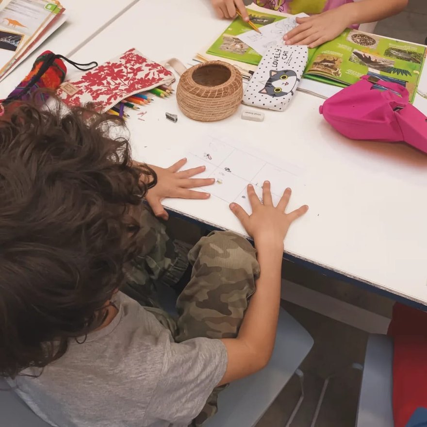 Chicos dibujando alrededor de una mesa en un taller, con lápices de colores, libros y una libreta abierta.