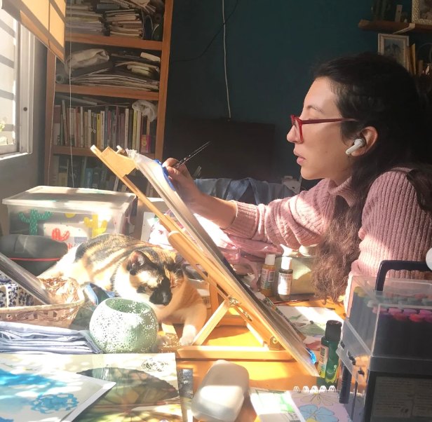 Tatiana pintando en su atril, con su gato sentado en el escritorio y la luz de la ventana entrando de costado.